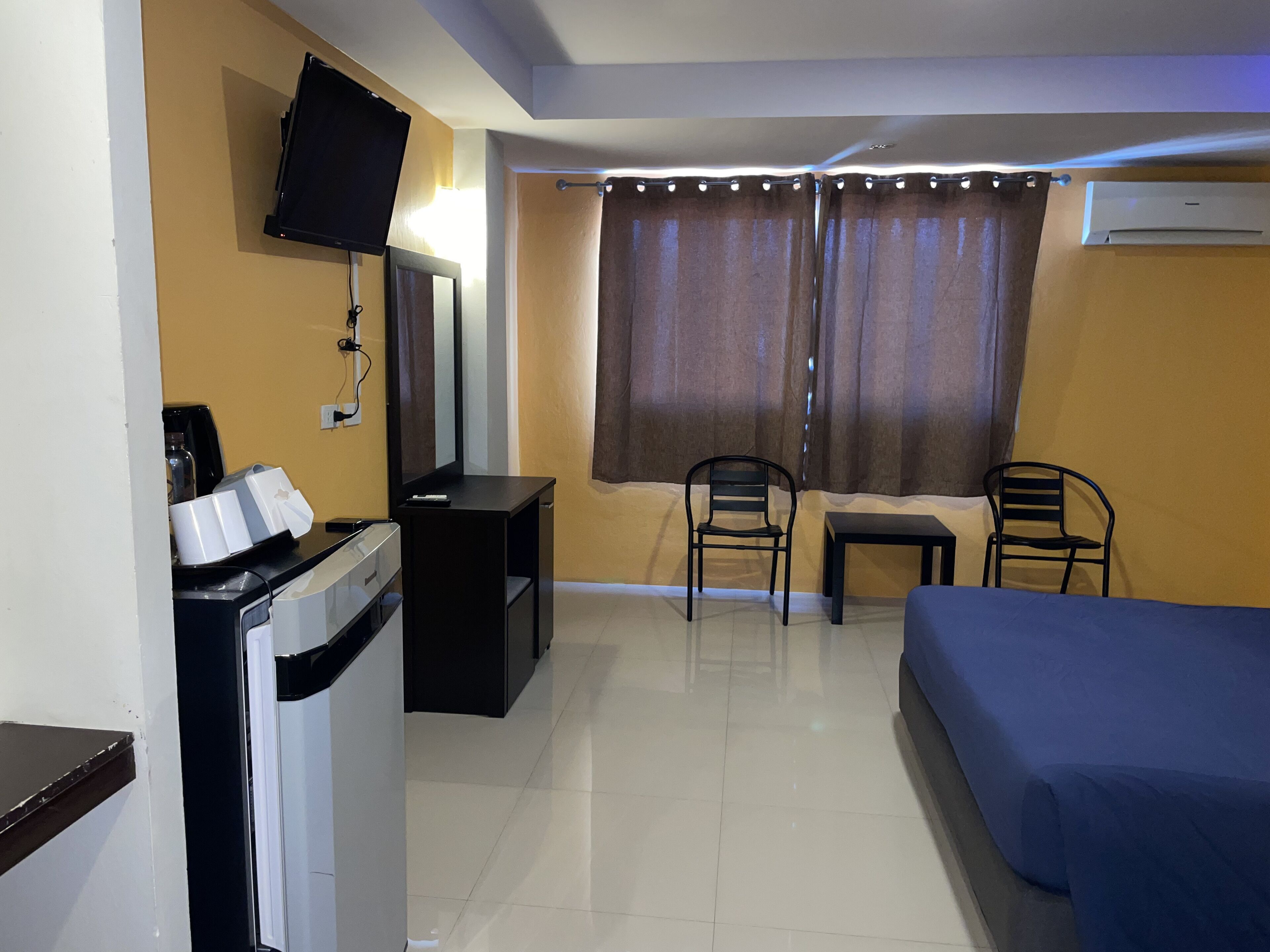 A hotel room with a bed, a desk with a computer, a chair, a TV mounted on the wall, and an air conditioning unit.