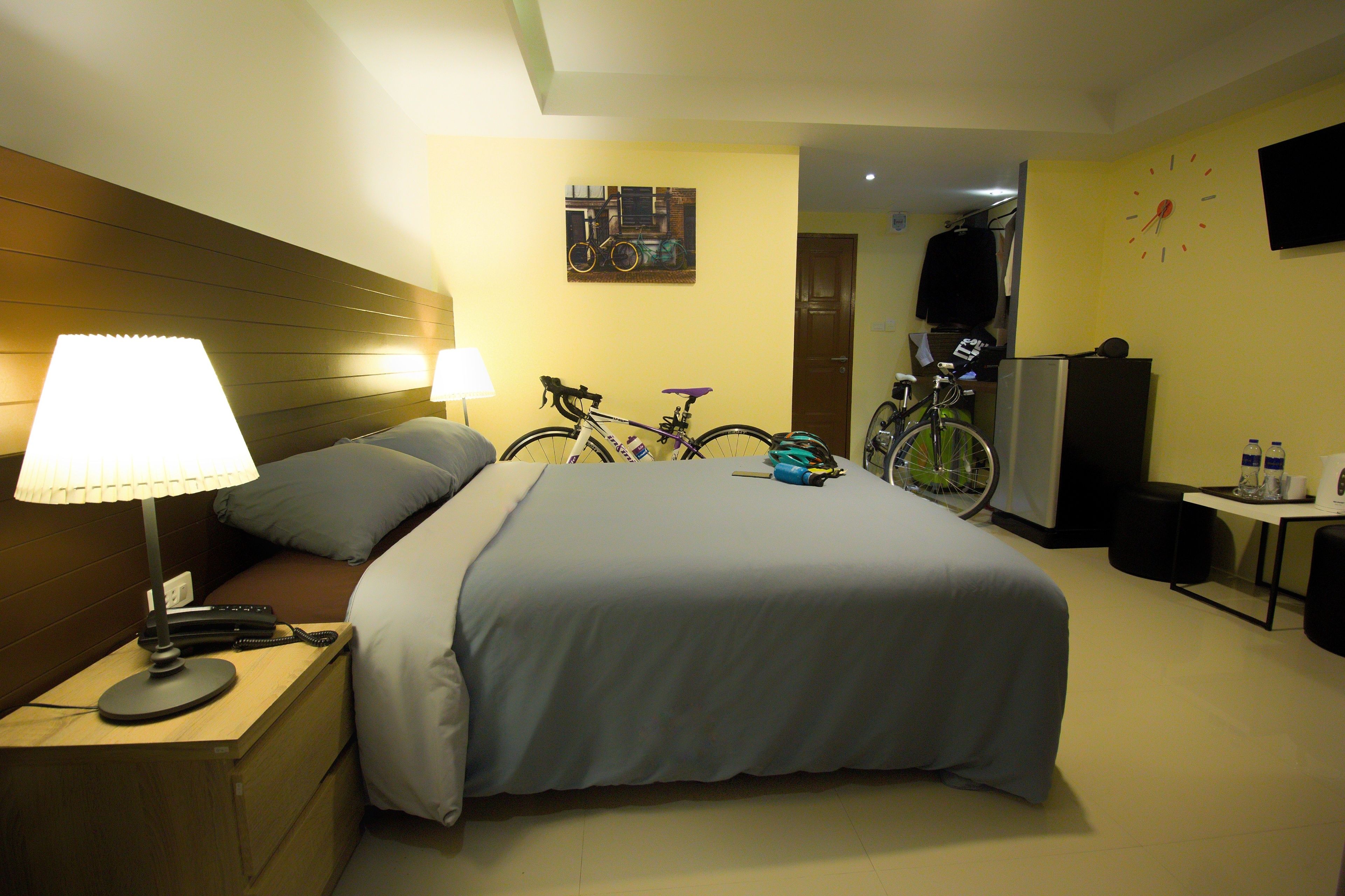 A hotel room with a bed, a nightstand with a lamp, a bicycle, a TV, and a clock on the wall.