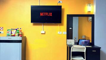 A room with a television mounted on the wall displaying 'NETFLIX', a small refrigerator, a desk with a laptop, a chair, and a mirror.