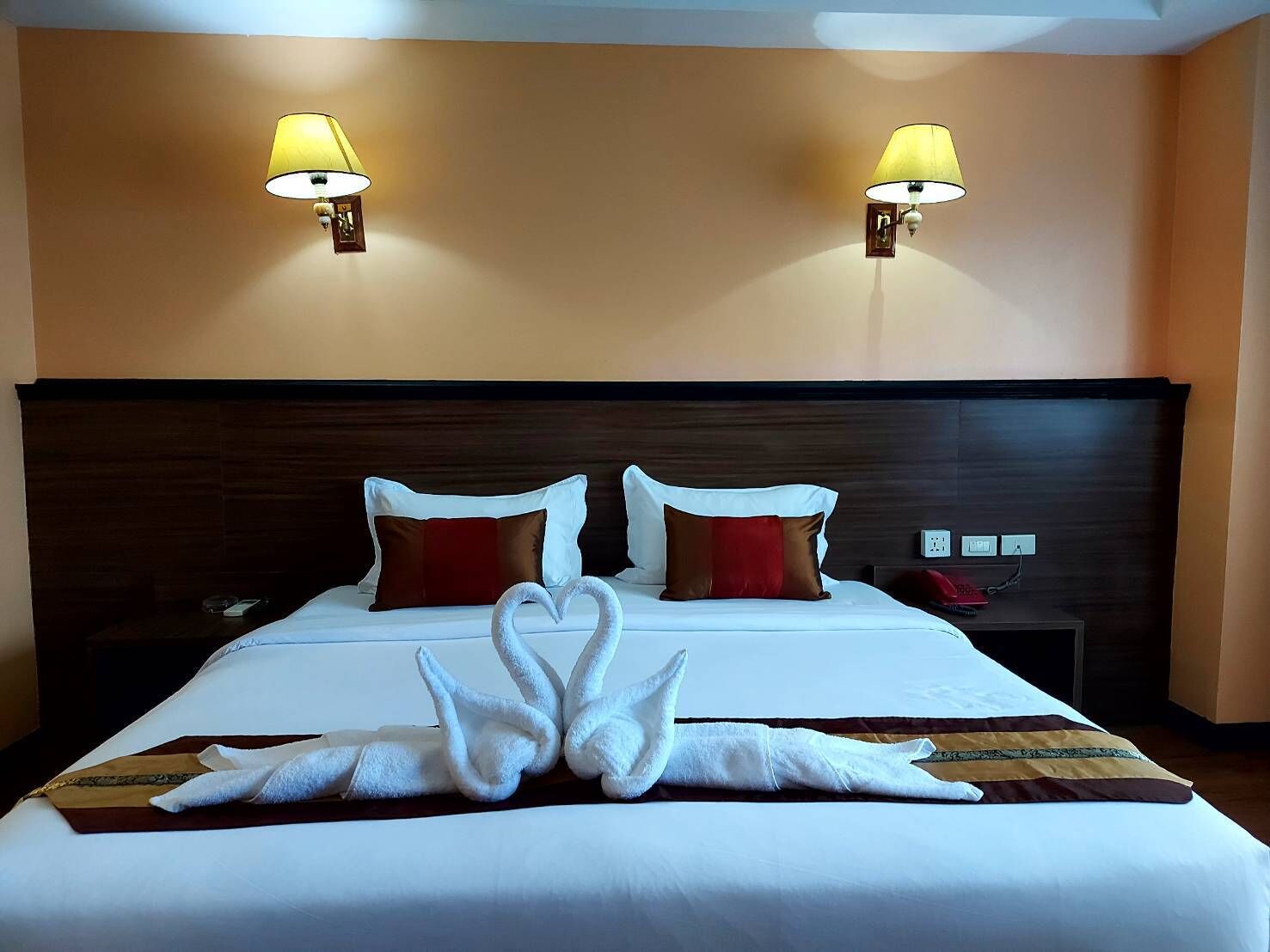 A hotel room with a large bed, two wall-mounted lamps, a wooden headboard, and a red and brown pillow.