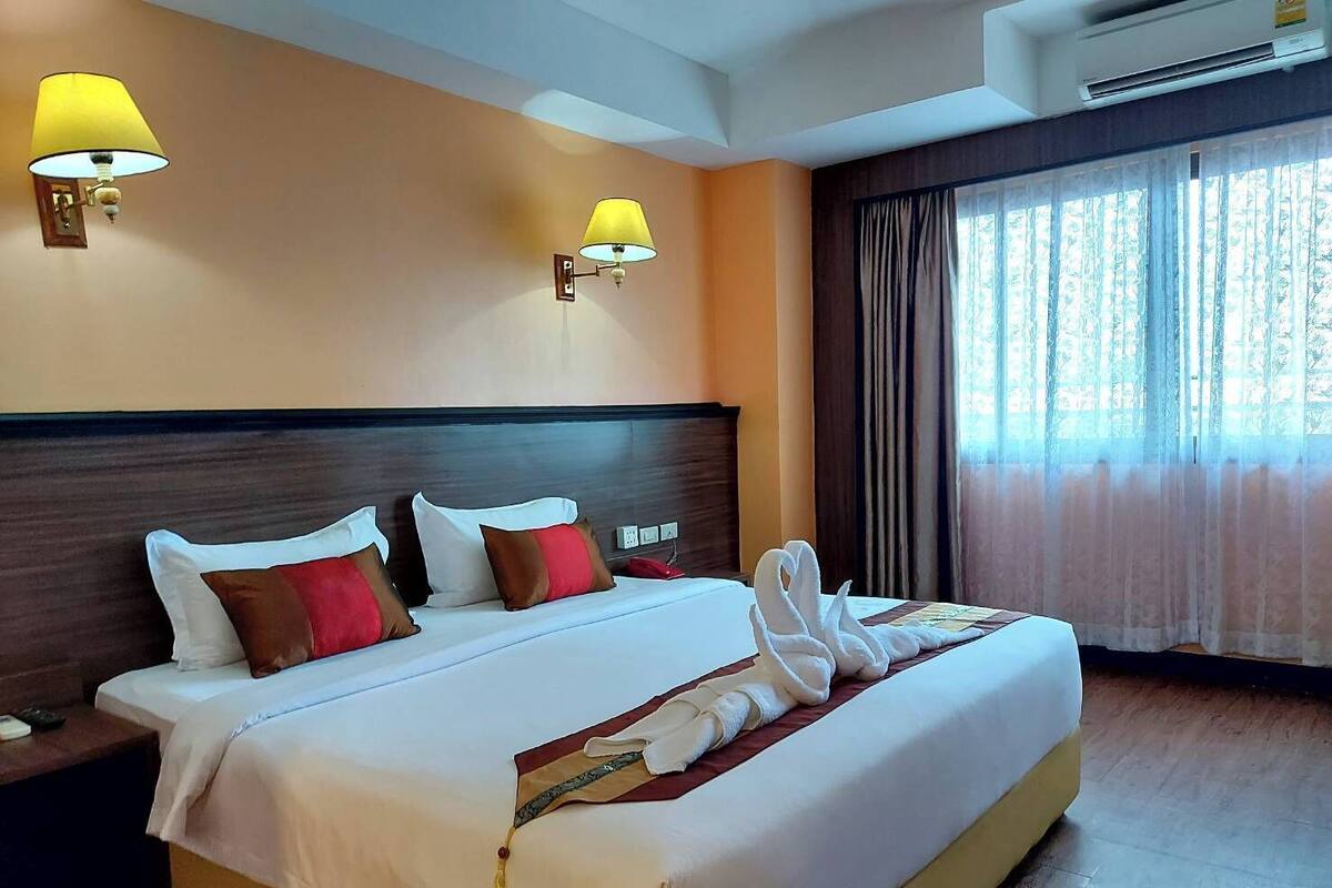 A hotel room with a large bed, two wall-mounted lamps, a window with curtains, and an air conditioning unit.