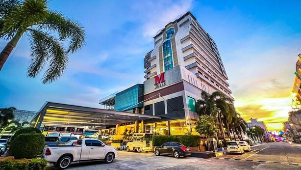 A modern hotel with a prominent 'M' logo, a glass entrance, and a parking lot with several cars. - M Hotel Danok (Sadao)