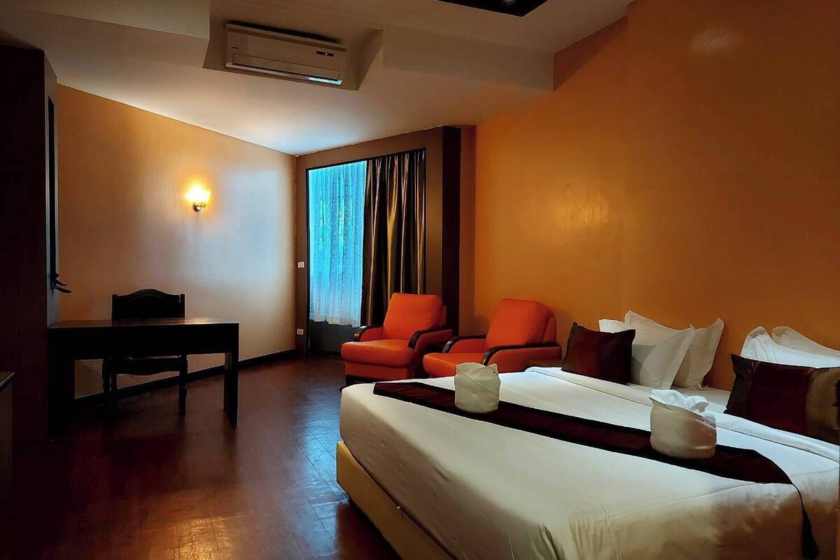 A hotel room with a bed, two orange chairs, a table, and a window with curtains.