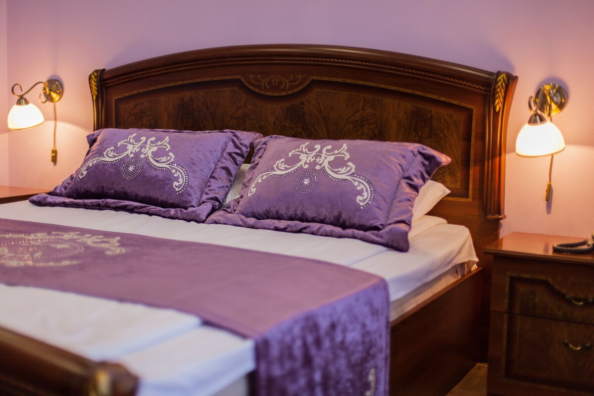 A bed with a wooden headboard, two purple pillows with decorative patterns, a white bedspread, and two bedside lamps on either side.