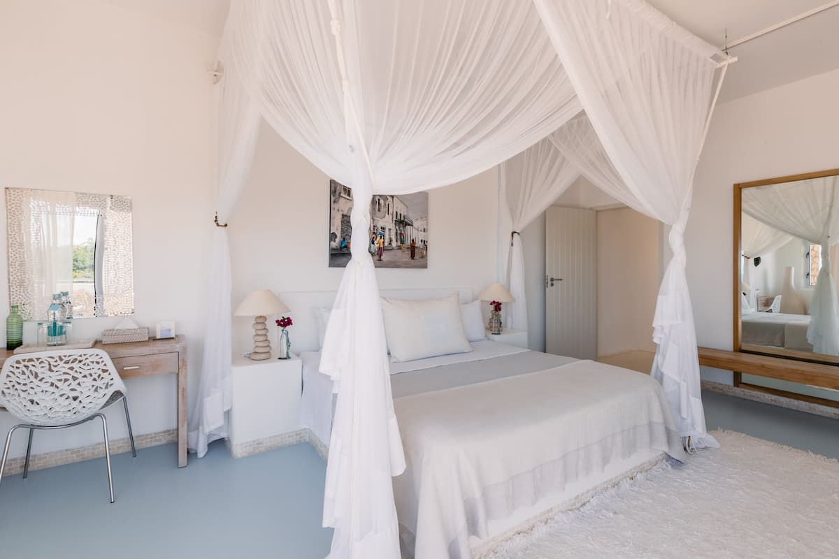 A bedroom with a four-poster canopy bed, a bedside table with a lamp, a chair, and a large mirror.