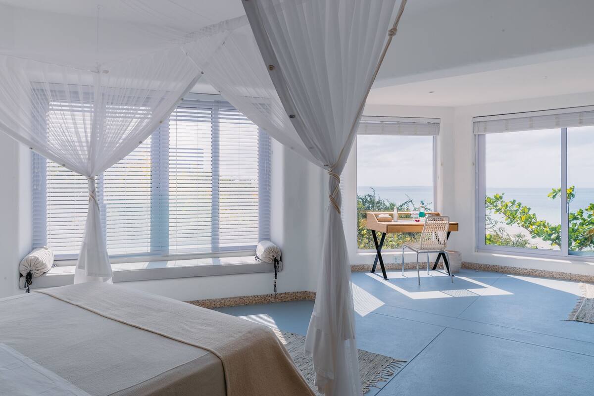 A bedroom with a bed, a desk, and large windows with a view of the ocean.