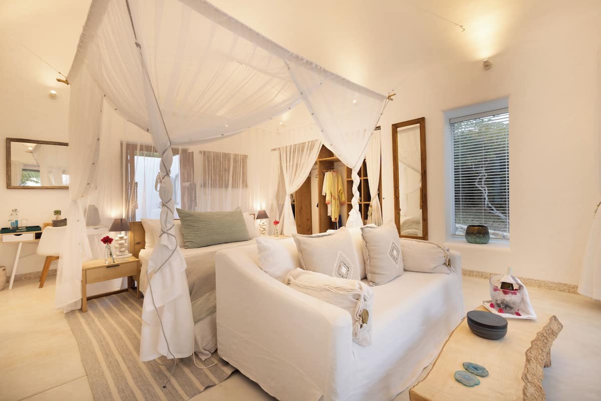 A bedroom with a canopy bed, a bedside table, a mirror, and a window with blinds.