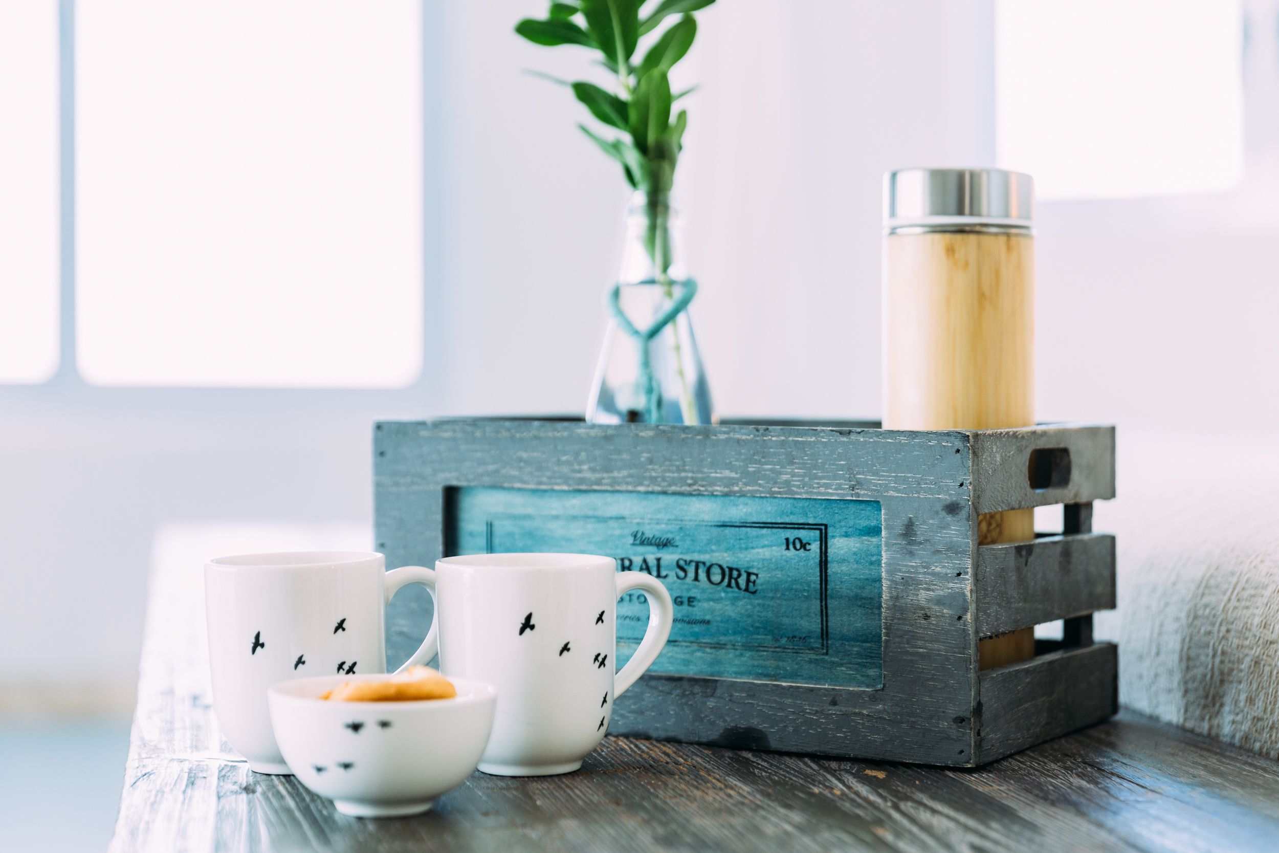 A rustic wooden crate with a vintage store sign, two white mugs with bird designs, a small bowl, and a thermos on a wooden surface.