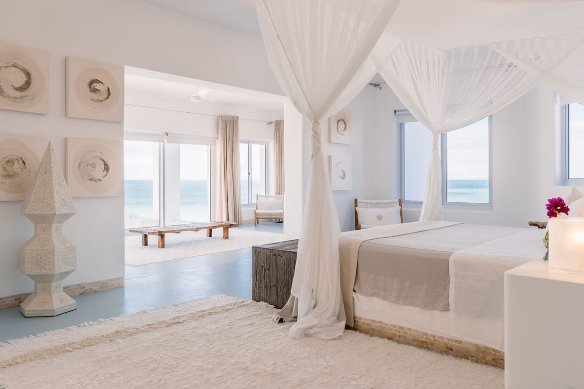 A bedroom with a large bed, a canopy, a view of the beach, and a white decor theme.