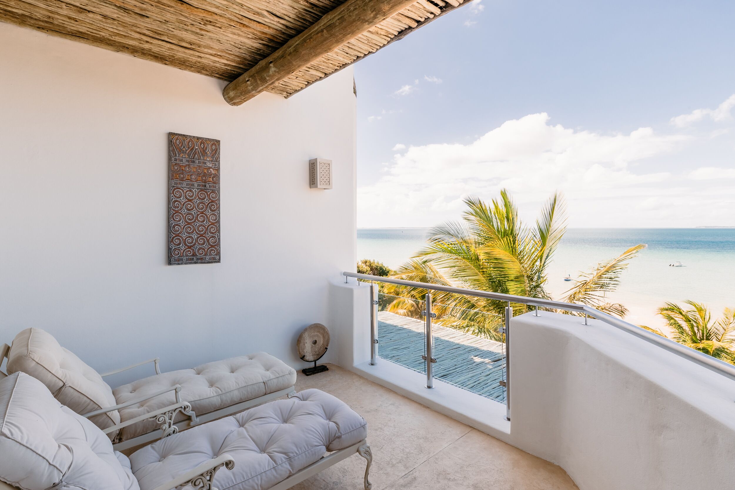 A balcony with a view of the beach, a cushioned sofa, a wall-mounted artwork, and a ceiling fan.