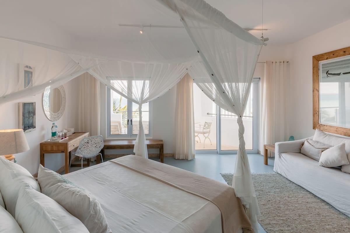 A spacious bedroom with a large bed, a desk, and a chair. There is a balcony with a view of the sea.