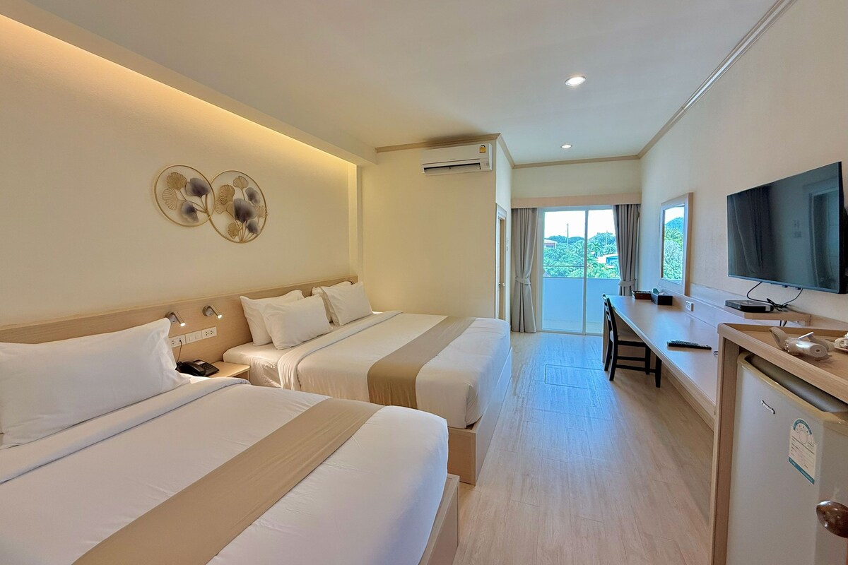 A hotel room with two beds, a desk, a television, and a view of greenery outside.