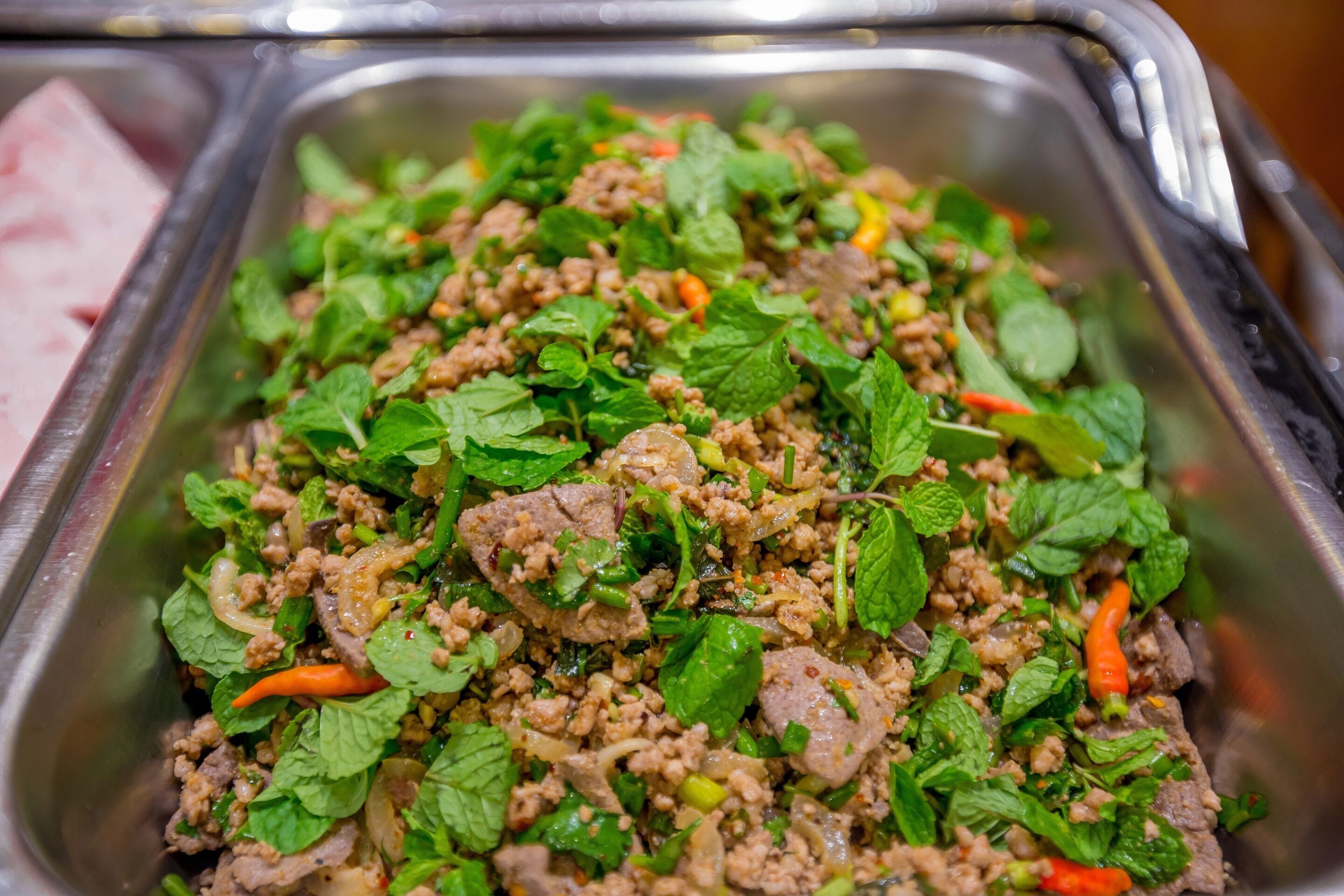 A stainless steel food serving tray filled with a dish of minced meat, herbs, and vegetables.