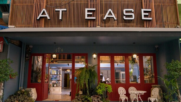 A storefront with the name "AT EASE" in large letters. There are potted plants and white chairs outside the entrance. - At Ease Bijou Place (Chiang Mai)