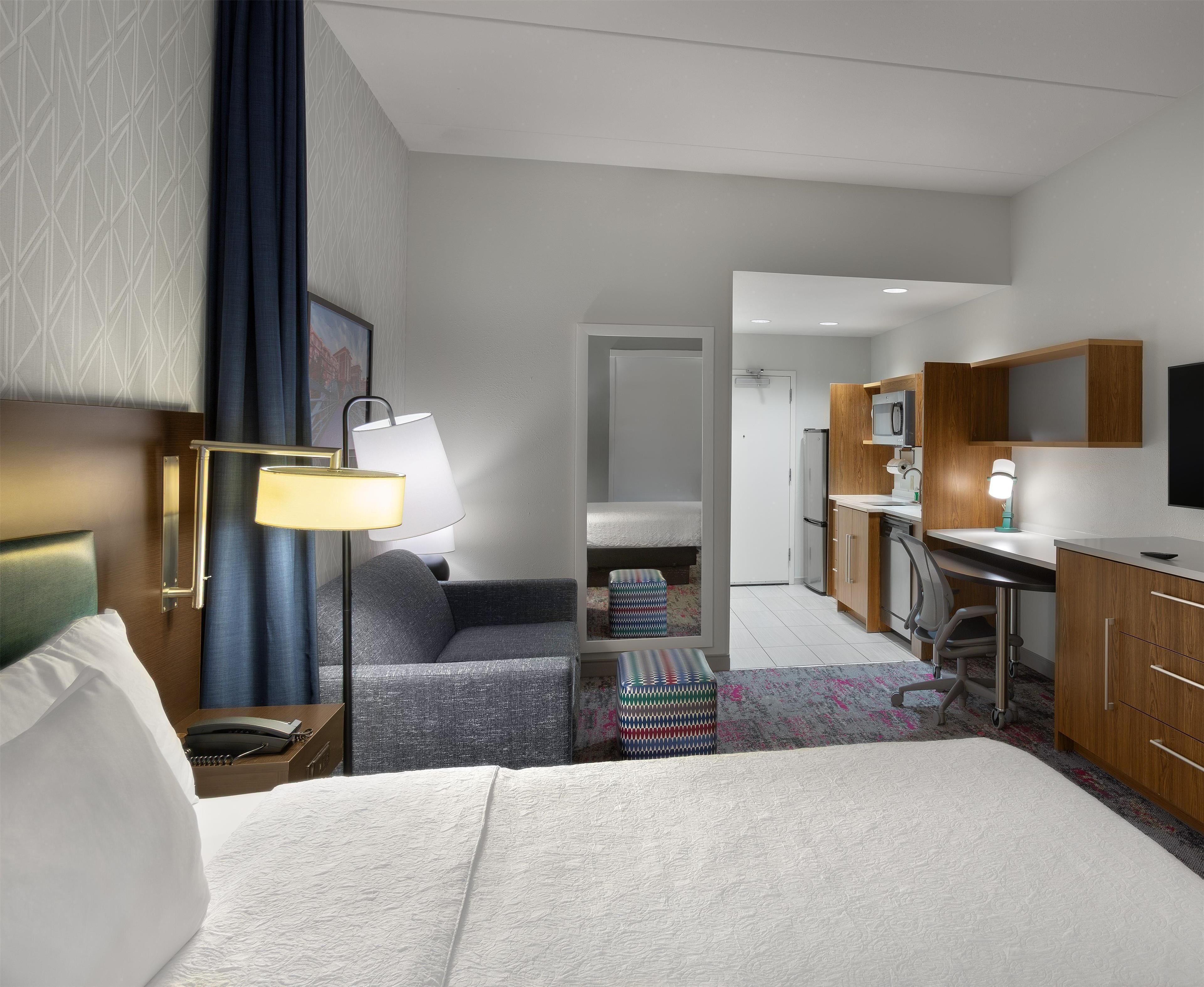 A modern hotel room with a bed, a desk, a chair, a lamp, a small table, and a kitchenette.