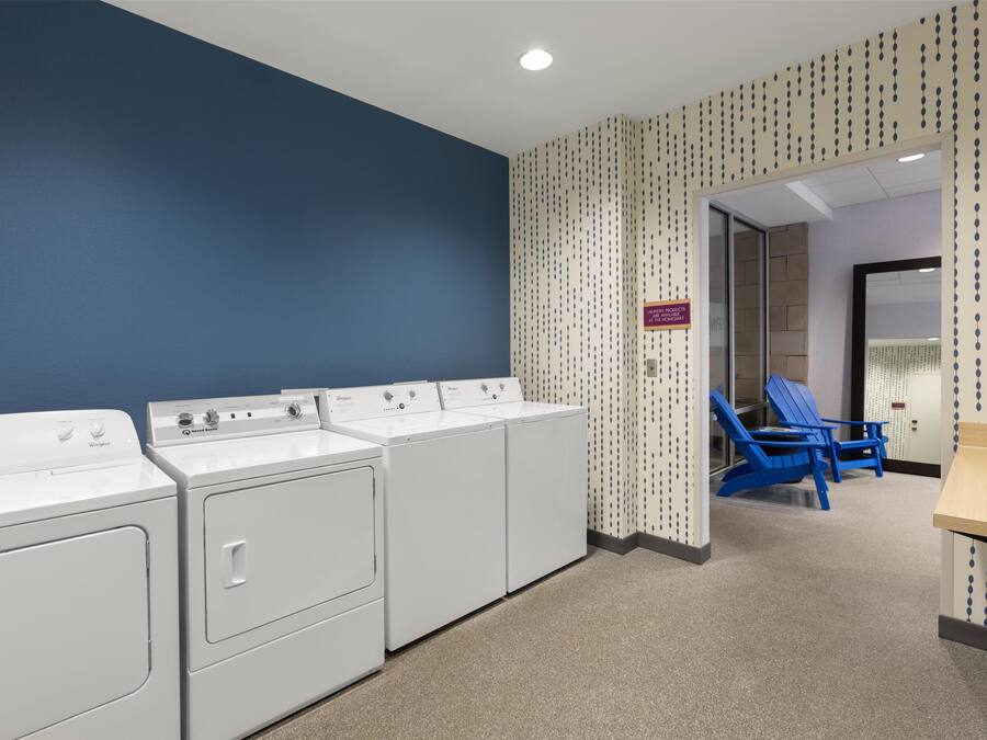 Una sala de lavandería con cuatro lavadoras y secadoras blancas, una pared azul, papel tapiz estampado y una silla azul al fondo.