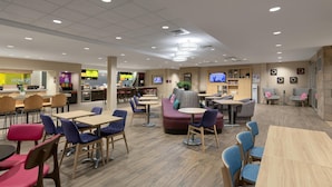 A modern hotel lobby with a central seating area, a kitchenette, and a TV screen. - Home2 Suites by Hilton Greenville Airport (Greenville)