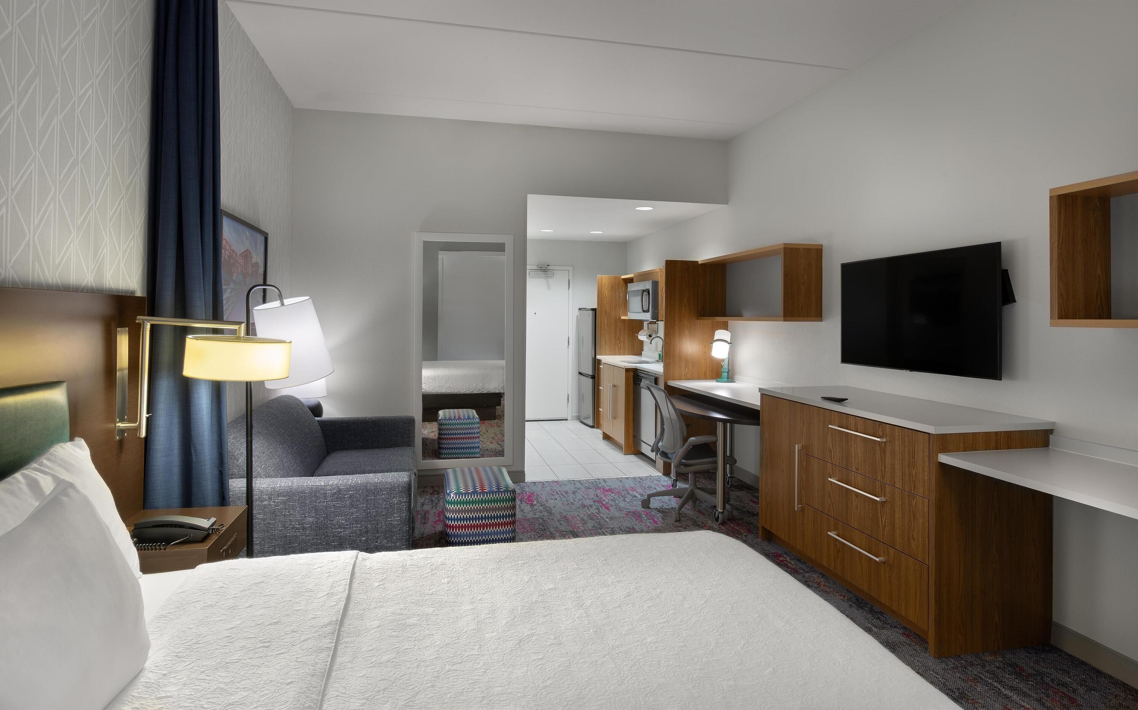 A hotel room with a bed, bedside table, lamp, desk, chair, kitchenette, and a TV.