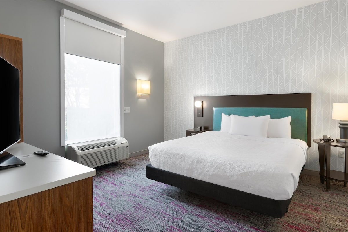 A hotel room with a bed, a desk, a TV, a heater, a lamp, and a patterned carpet.