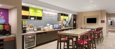 A modern hotel lobby with a self-service coffee station, an 'inspired table' area, and a TV mounted on the wall.