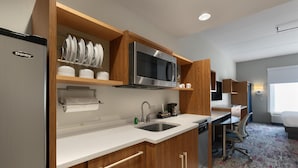 A modern kitchen with a stainless steel refrigerator, microwave, and sink. There are wooden cabinets and a countertop. A small office area with a desk and chair is visible in the background. - Home2 Suites by Hilton Greenville Airport (Greenville)