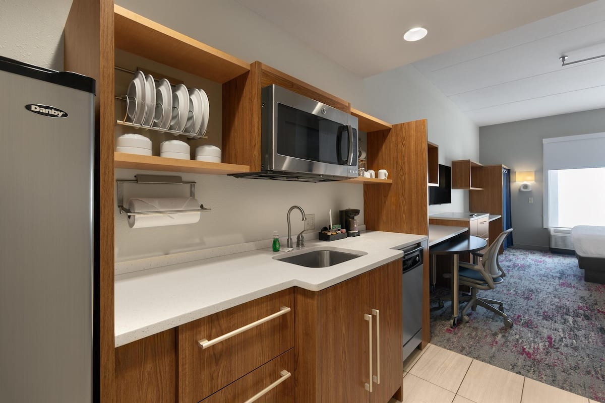 A modern kitchen with a stainless steel refrigerator, microwave, and sink. There are wooden cabinets and a countertop. A small office area with a desk and chair is visible in the background.