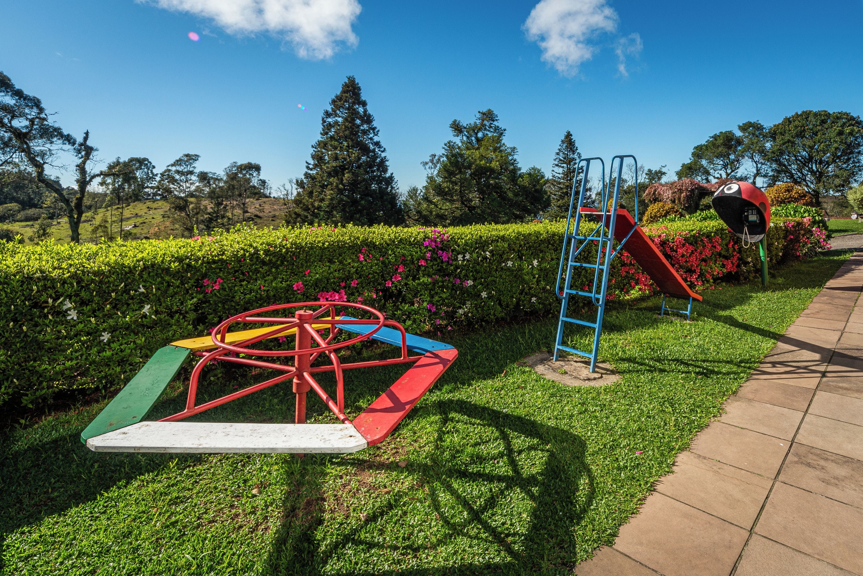 Un parque infantil con un tobogán y una estructura de escalada, rodeado de setos cuidados y plantas con flores.