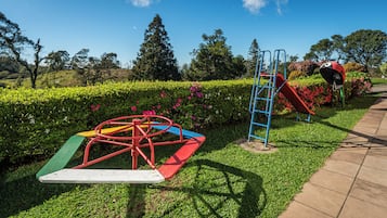 Un parque infantil con un tobogán y una estructura de escalada, rodeado de setos cuidados y plantas con flores.
