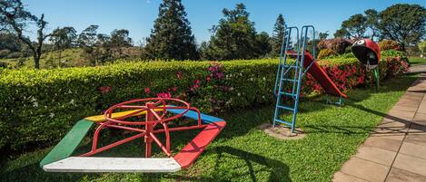 Un parque infantil con un tobogán y una estructura de escalada, rodeado de setos cuidados y plantas con flores.