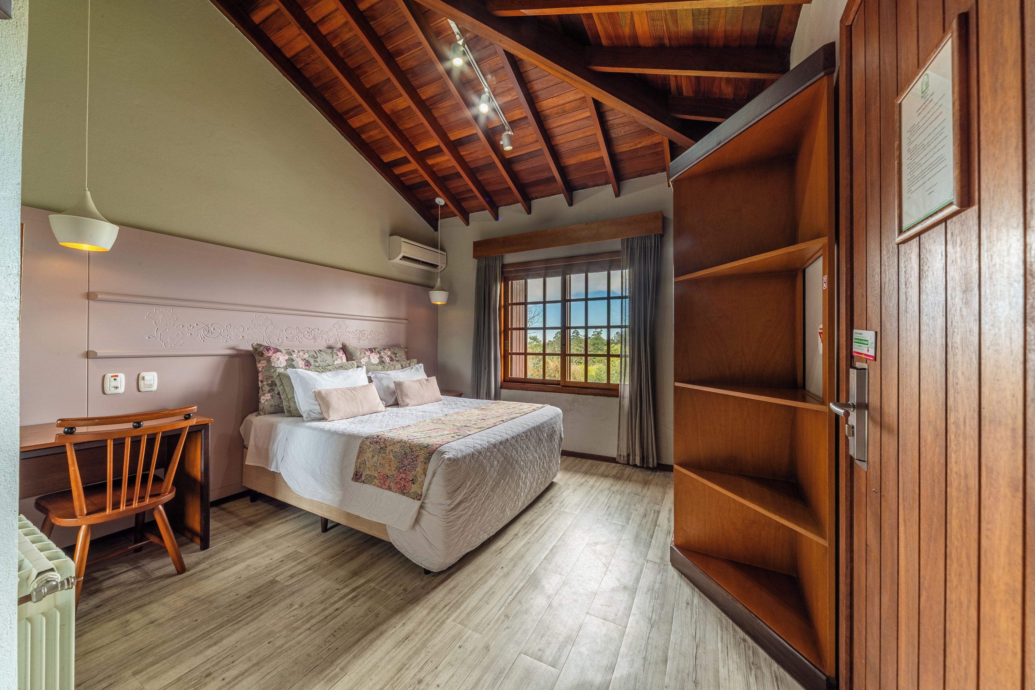 A bedroom with a wooden ceiling, a bed with floral bedding, a wooden chair, and a window with a view.