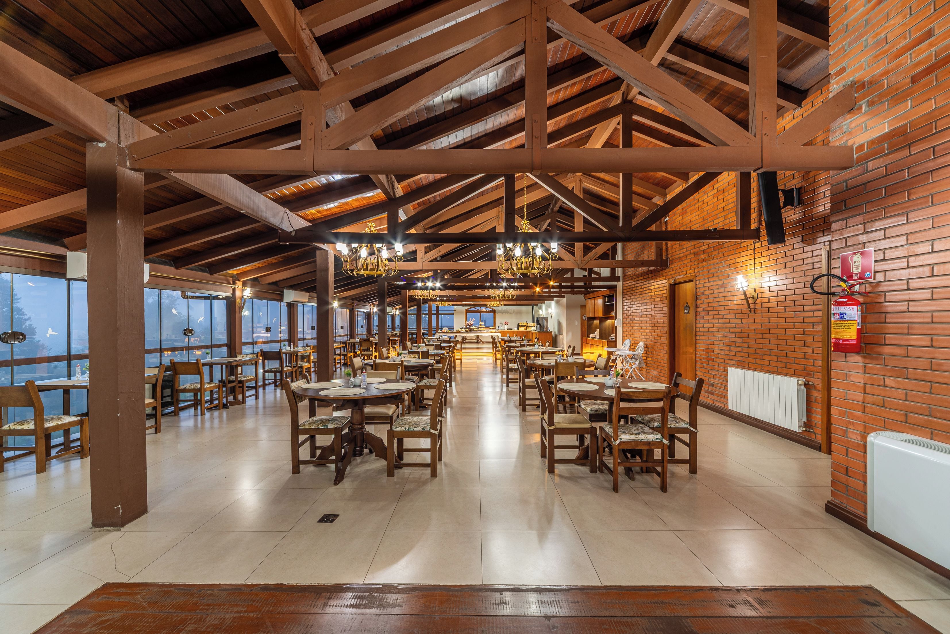 A spacious restaurant with high ceilings, exposed wooden beams, and brick walls. Tables and chairs are arranged for dining.
