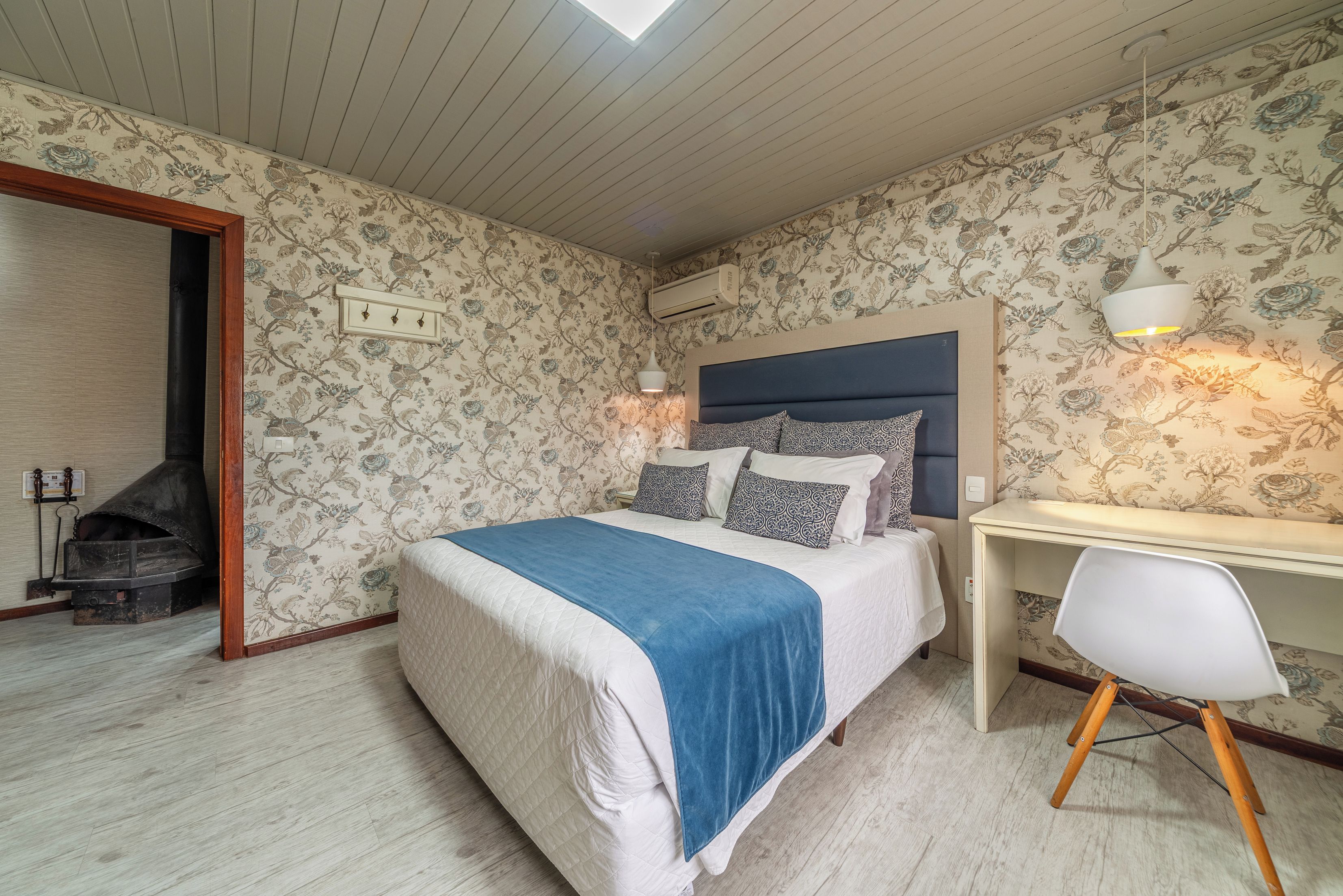 A bedroom with a bed, a desk, a chair, a fireplace, and floral wallpaper.