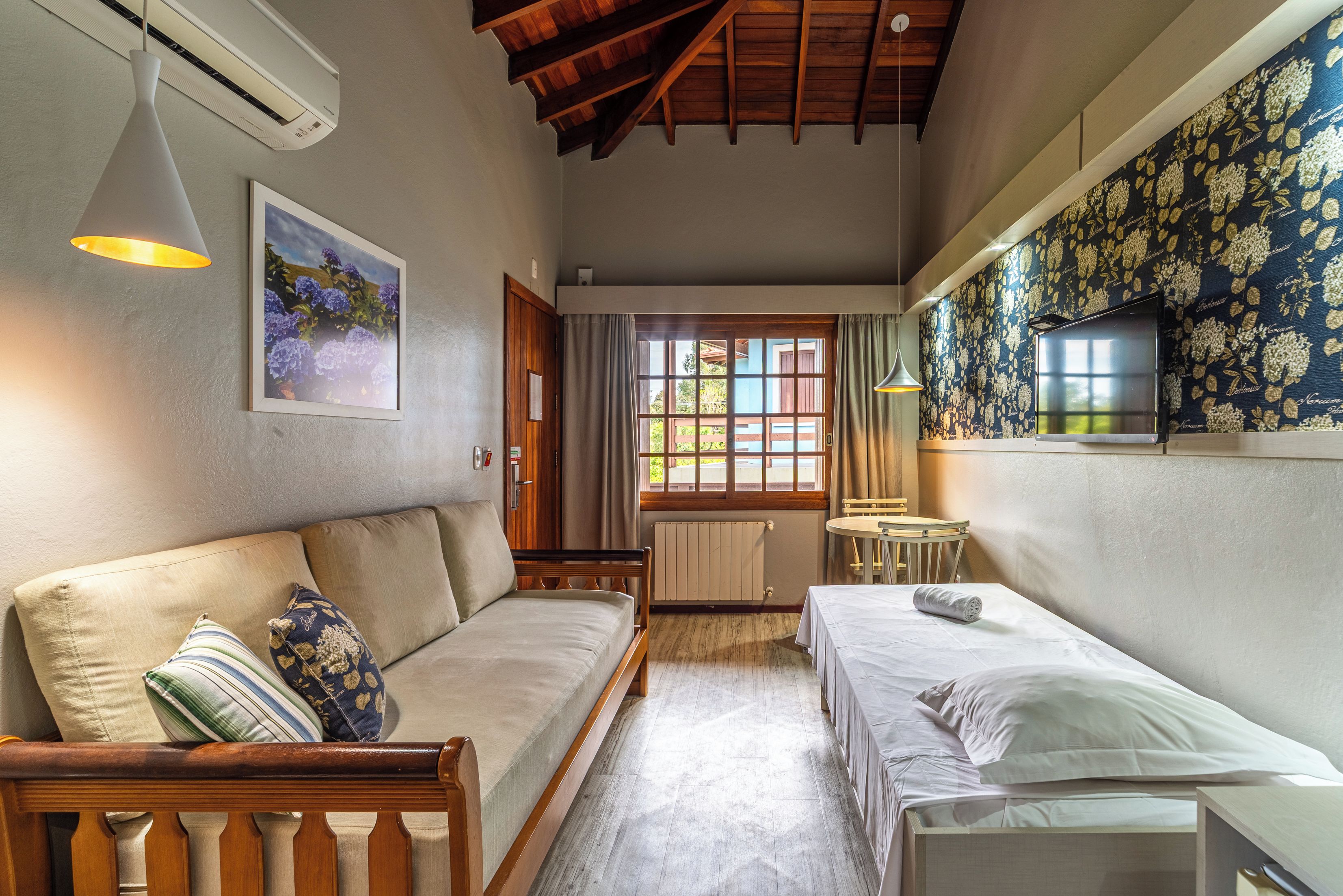A room with a wooden ceiling, a sofa, a bed, a small table, and a wall-mounted TV.