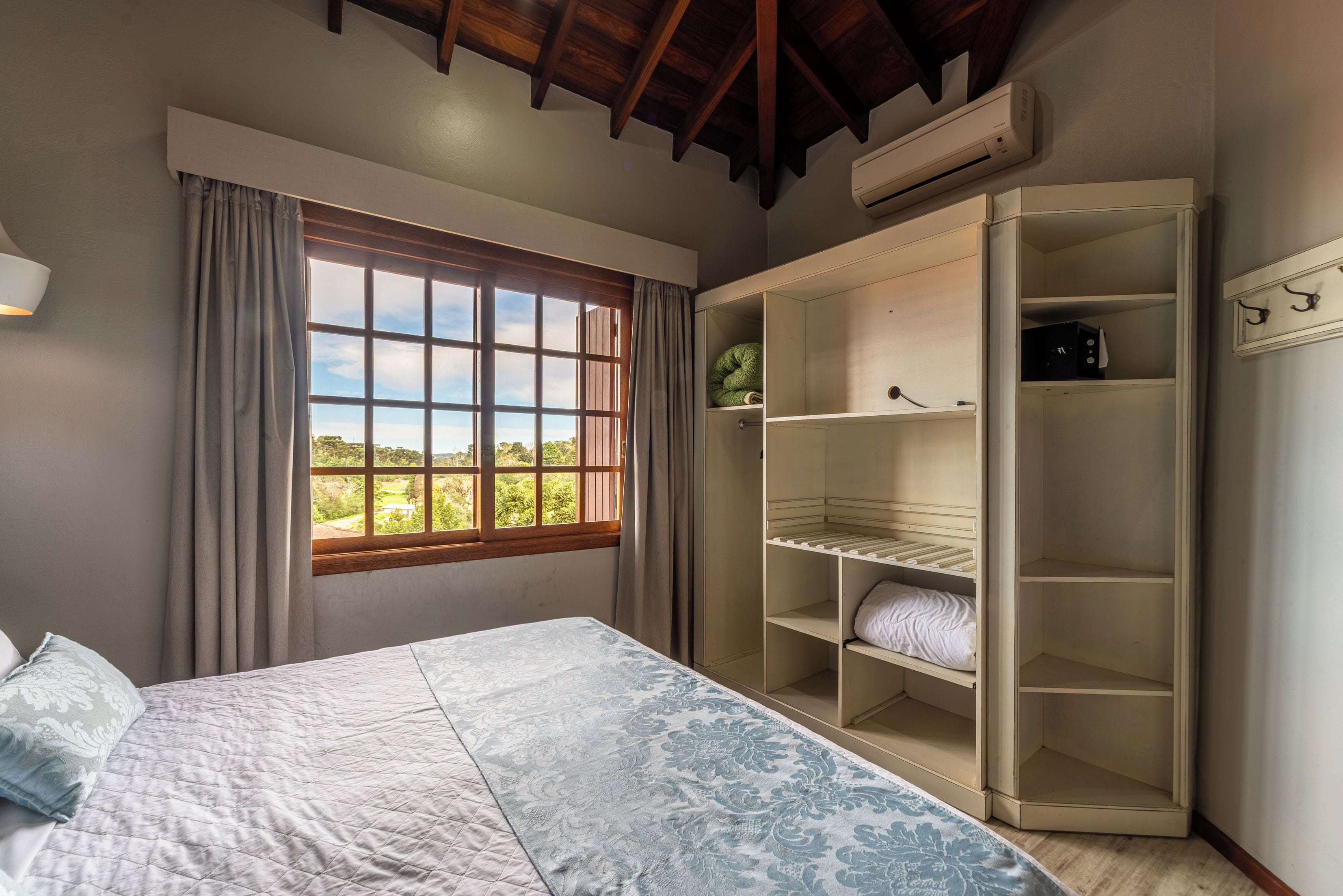 A bedroom with a bed, a window with curtains, a wardrobe with shelves, and an air conditioning unit.