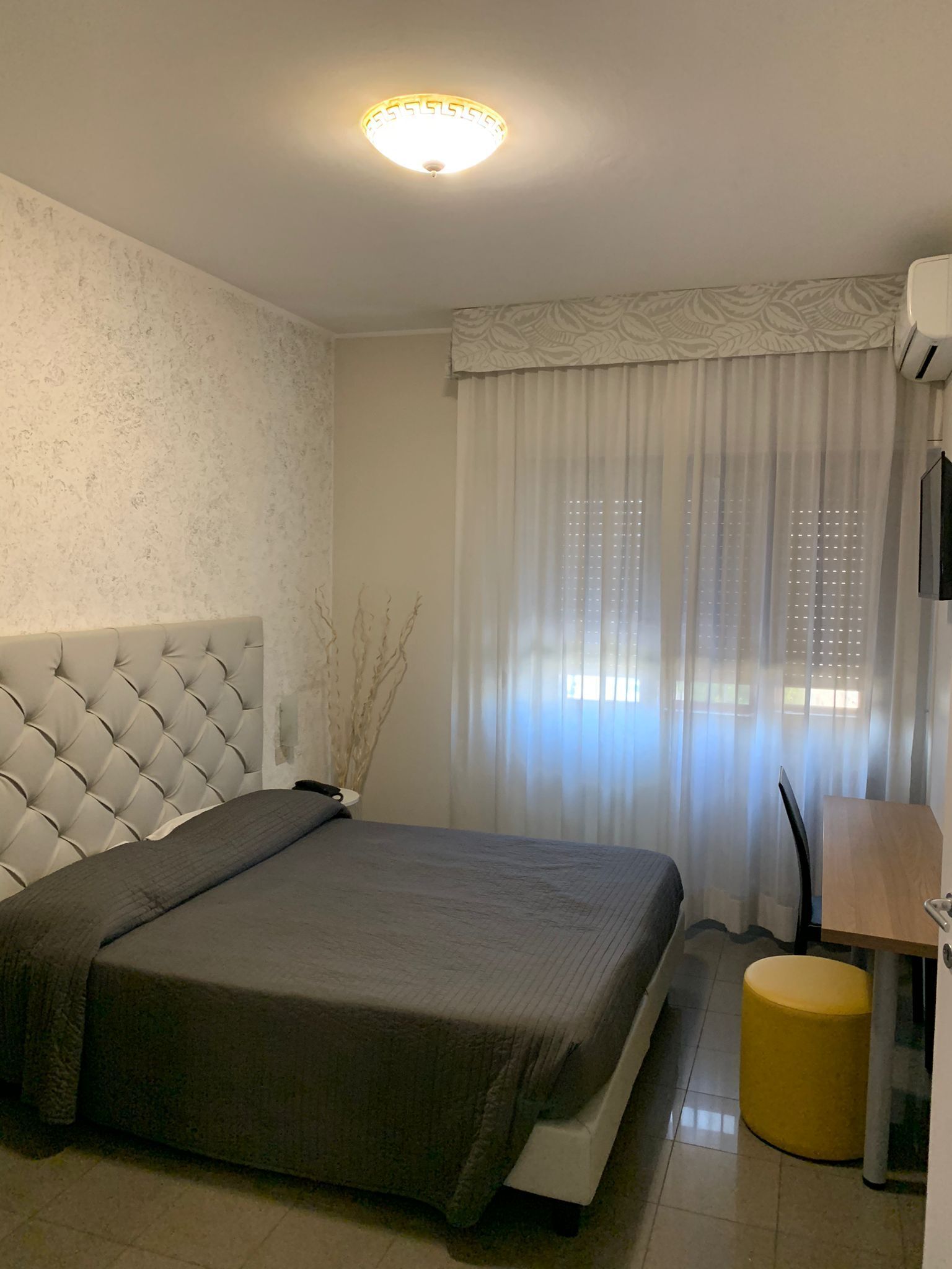 A modern hotel room with a tufted headboard, a large bed, a bedside table, a small stool, and a window with sheer curtains.