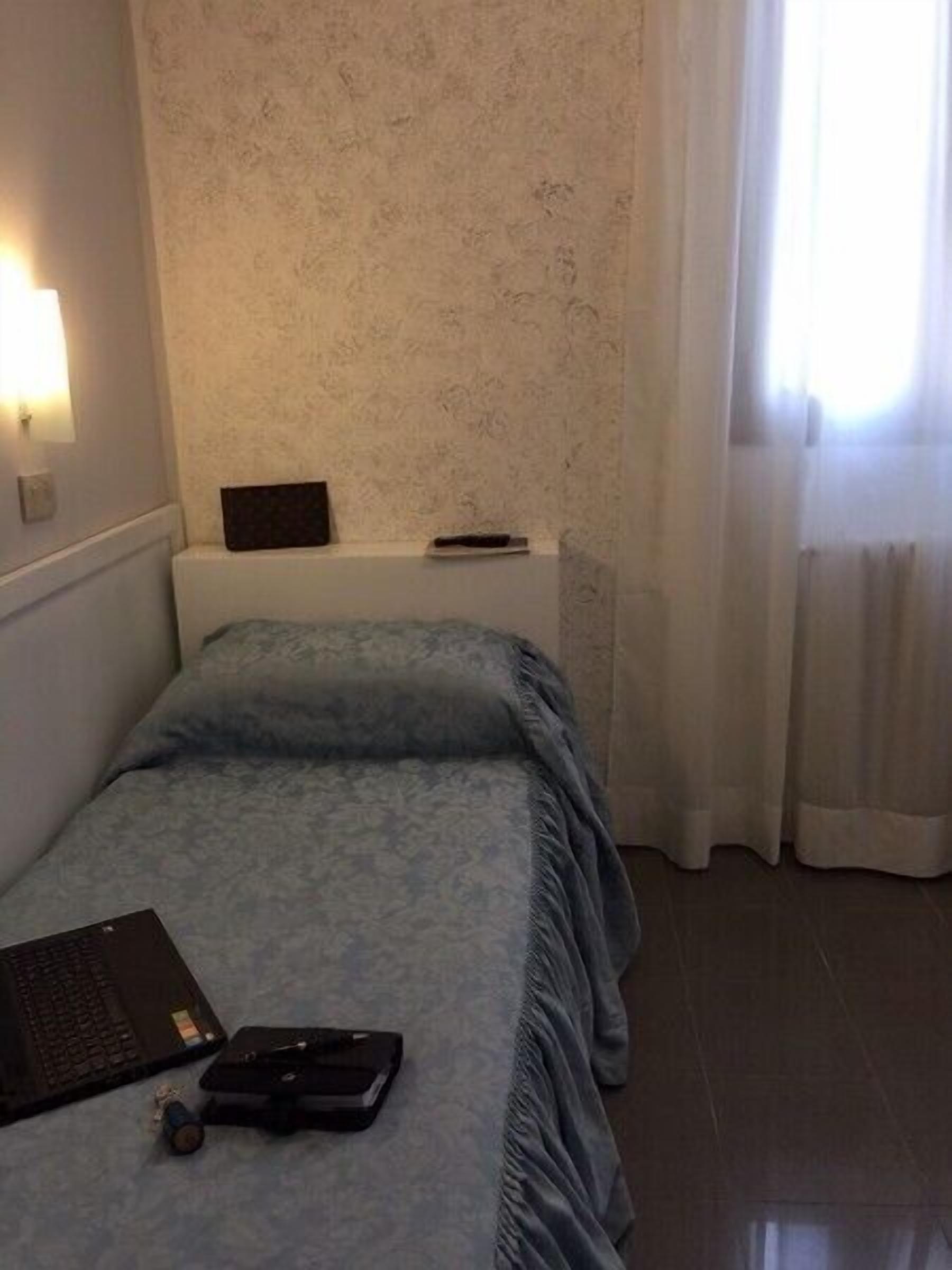 Ein kleines, ordentlich eingerichtetes Schlafzimmer mit Bett, Nachttisch, Lampe und einem Fenster mit Vorhängen.