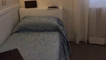 Ein kleines, ordentlich eingerichtetes Schlafzimmer mit Bett, Nachttisch, Lampe und einem Fenster mit Vorhängen.