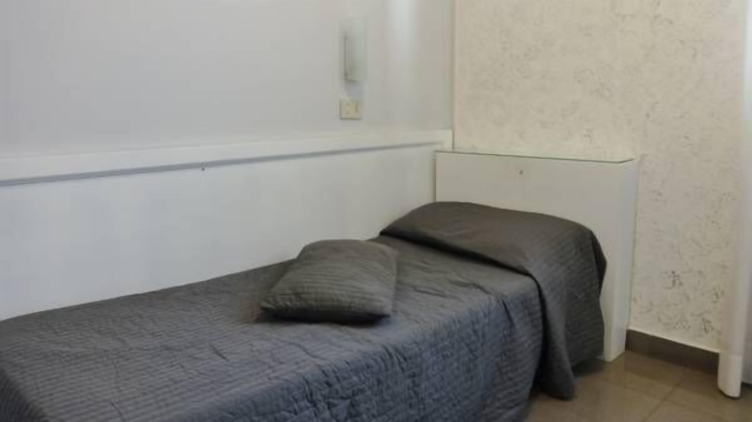 A small, single-bed room with a grey bedspread and pillow, a white wall, and a plain, light-colored floor.