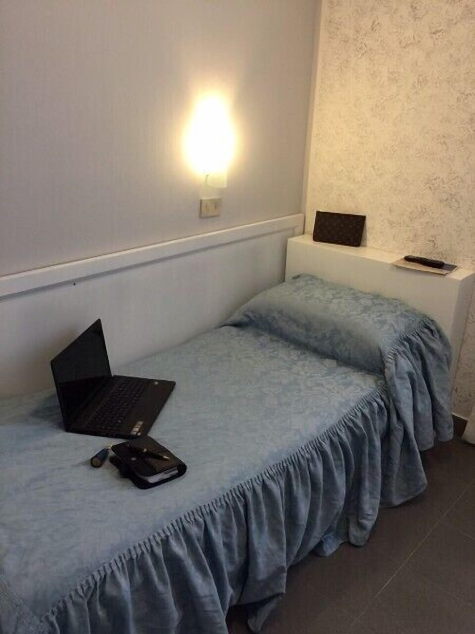 A small, single-bed room with a laptop and a remote control on the bed.