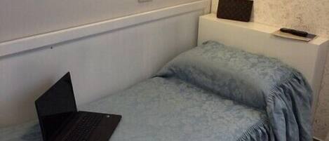 A small, single-bed room with a laptop and a remote control on the bed.
