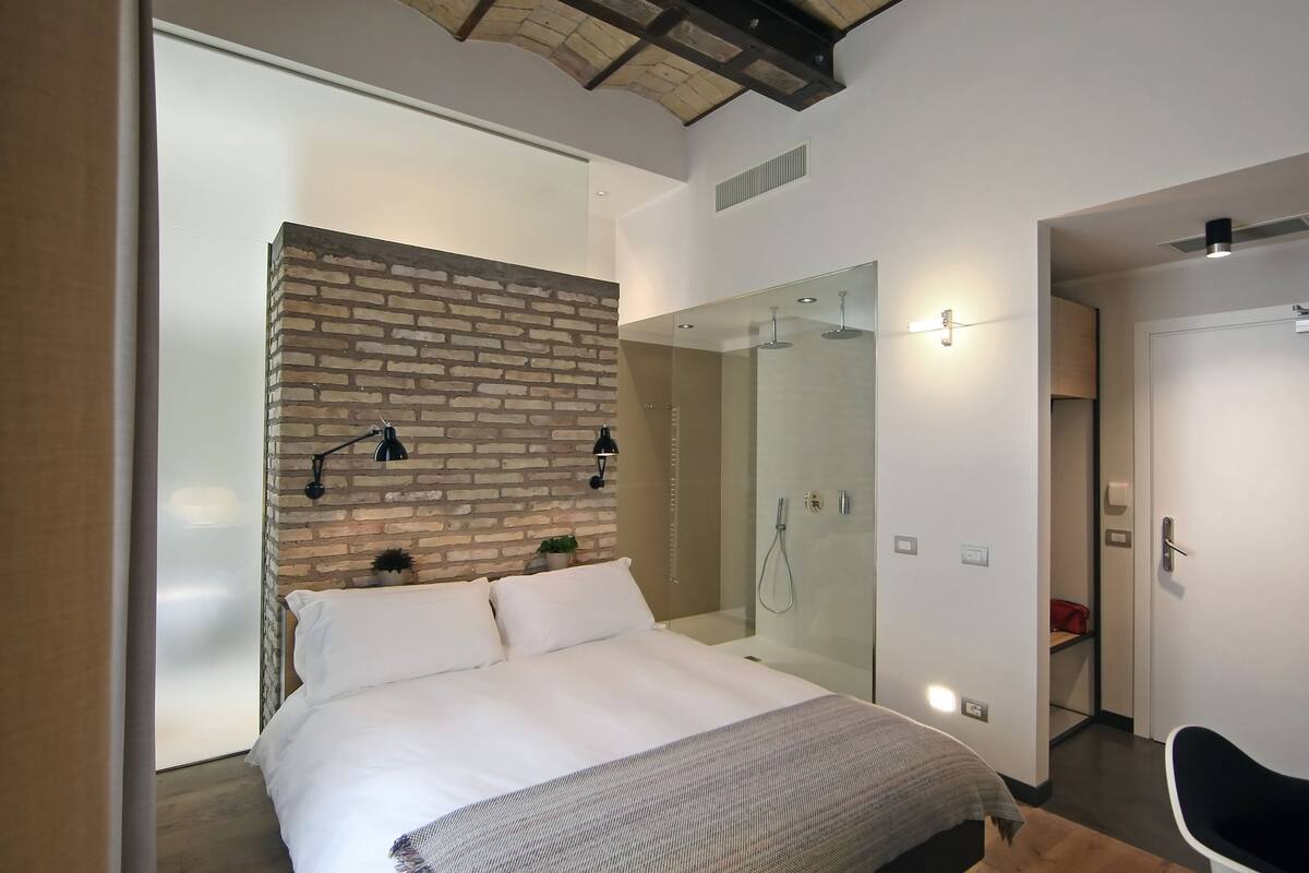 A modern bedroom with a brick accent wall, a large bed, a glass shower enclosure, and a minimalist design.