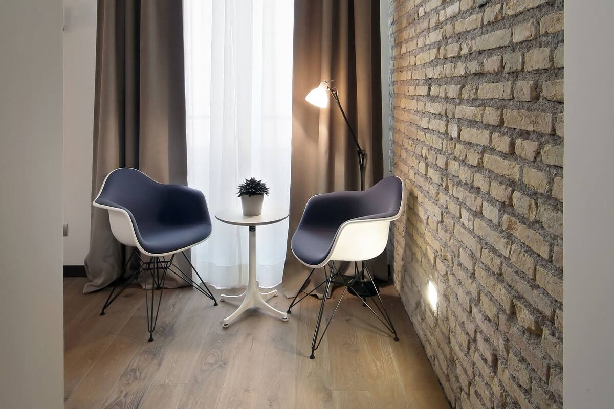 A room with a brick wall, two modern chairs, a small table, and a window with curtains.