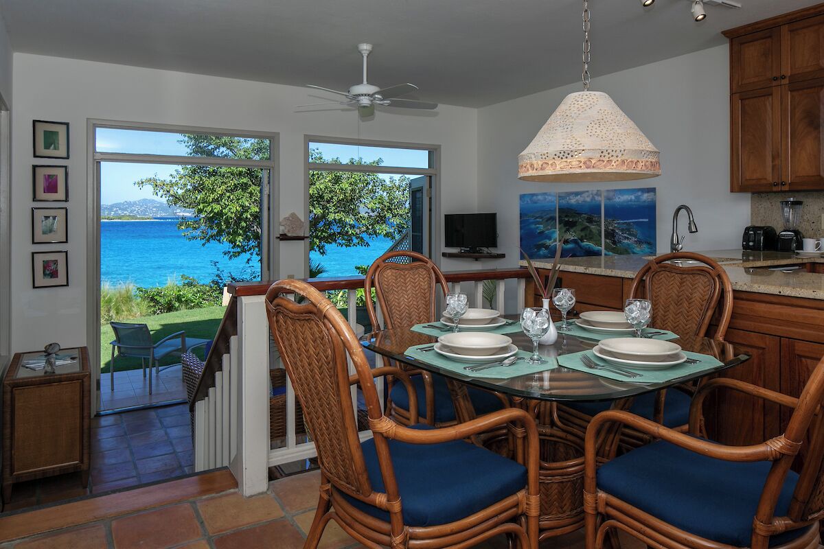 A dining area with a view of the sea, wicker chairs, a glass table, and a ceiling fan.