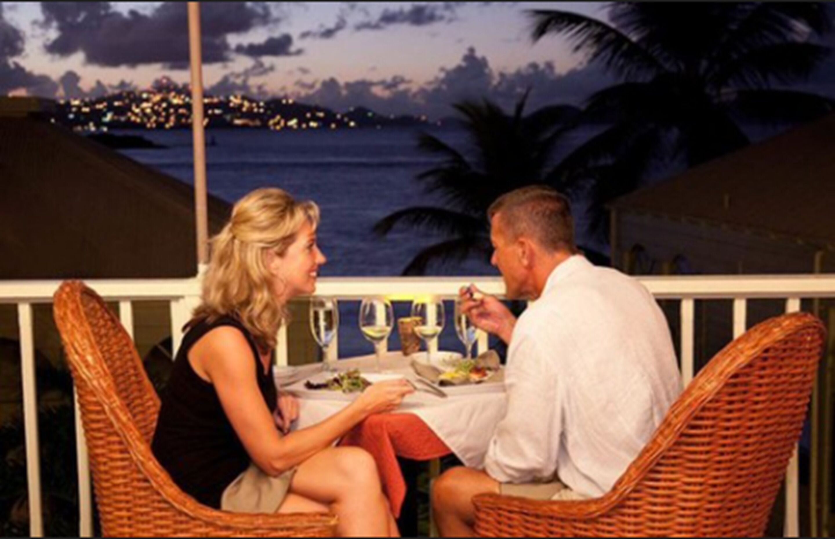 A couple dining at an outdoor table with a view of the ocean and city lights in the background.