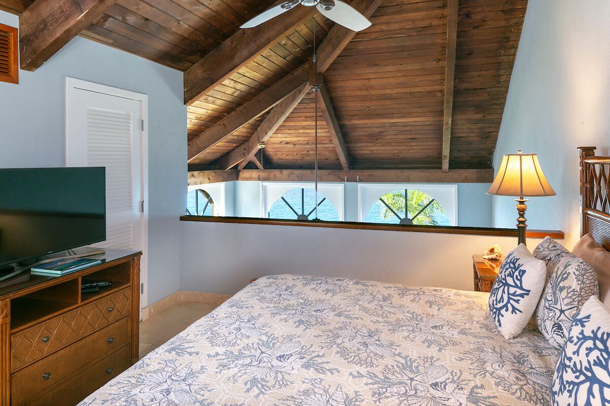 A bedroom with a sloped ceiling, a bed with floral bedding, a television on a wooden cabinet, and a window with a view of greenery.