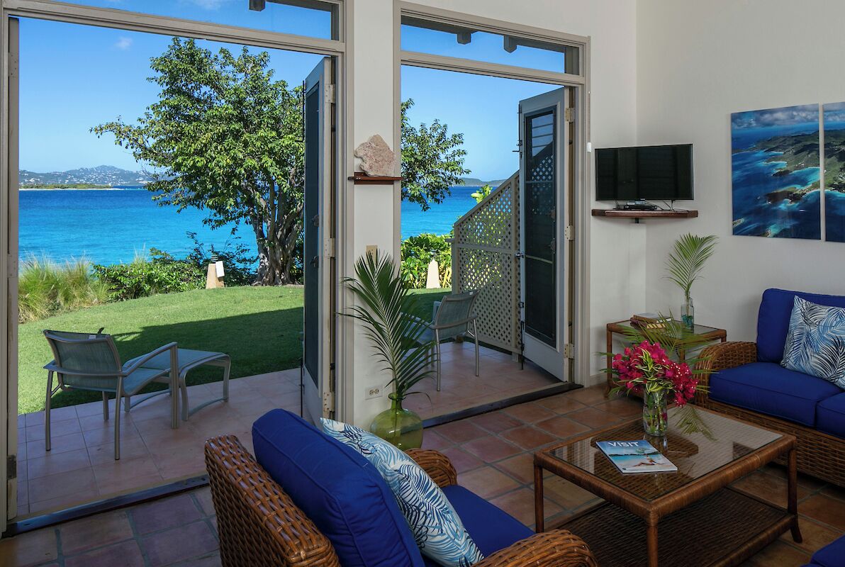 Un vaste espace de vie avec une grande porte vitrée donnant sur un balcon, un coin salon confortable avec un canapé et des fauteuils, un téléviseur à écran plat fixé au mur et une vue sur la mer.