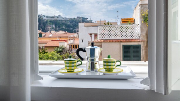 Vista desde la ventana de un paisaje urbano con edificios, cielo despejado y una mesa con una cafetera y tazas.