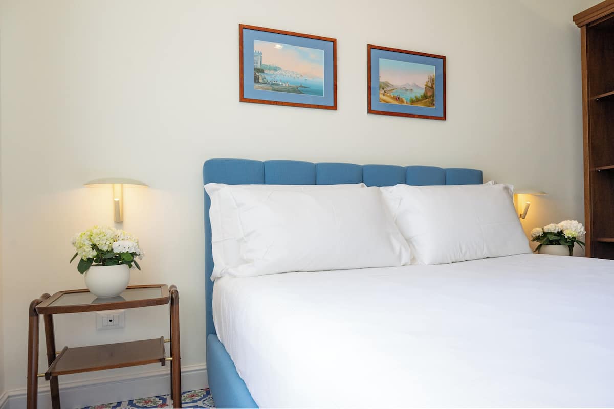 A neatly made bed with white linens, a blue headboard, and two wall-mounted paintings.