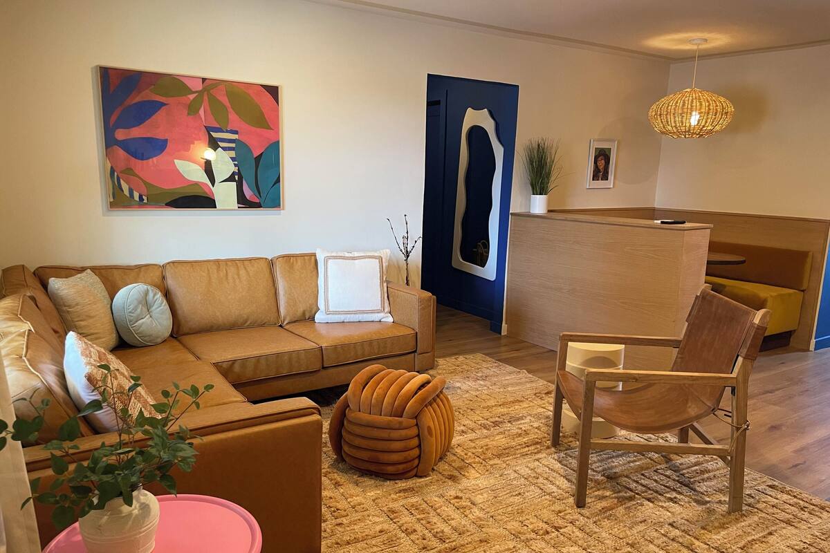 Ett modernt vardagsrum med en brun soffa, en trästol, ett rosa bord och en stor, abstrakt tavla på väggen.