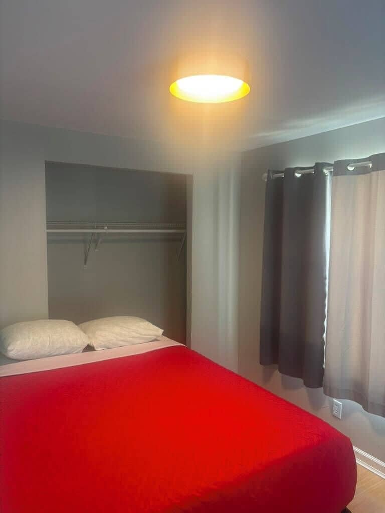 Ein Zimmer mit einem Bett, einer Pendelleuchte, einem Kleiderhaken und einem Fenster mit Vorhängen.
